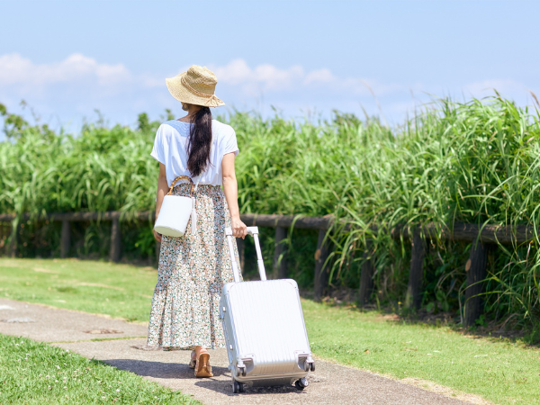 青空の下スーツケースを引いて歩く麦わら帽子をかぶったロングヘア&ロングスカートの女性の後ろ姿